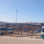 Bus terminal with several white and turquoise minibuses under sheltered bays; urban skyline in the distance.