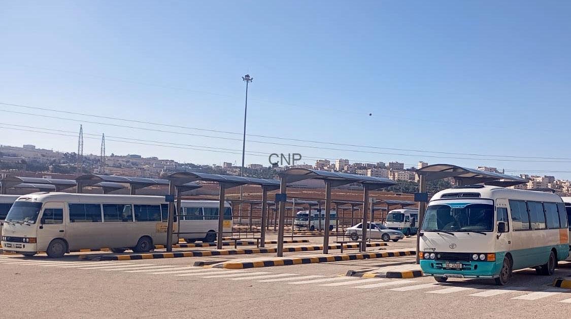 Bus terminal with several white and turquoise minibuses under sheltered bays; urban skyline in the distance.