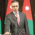 Man in a dark suit and pink tie speaks at a podium, with Jordanian flags in the background.