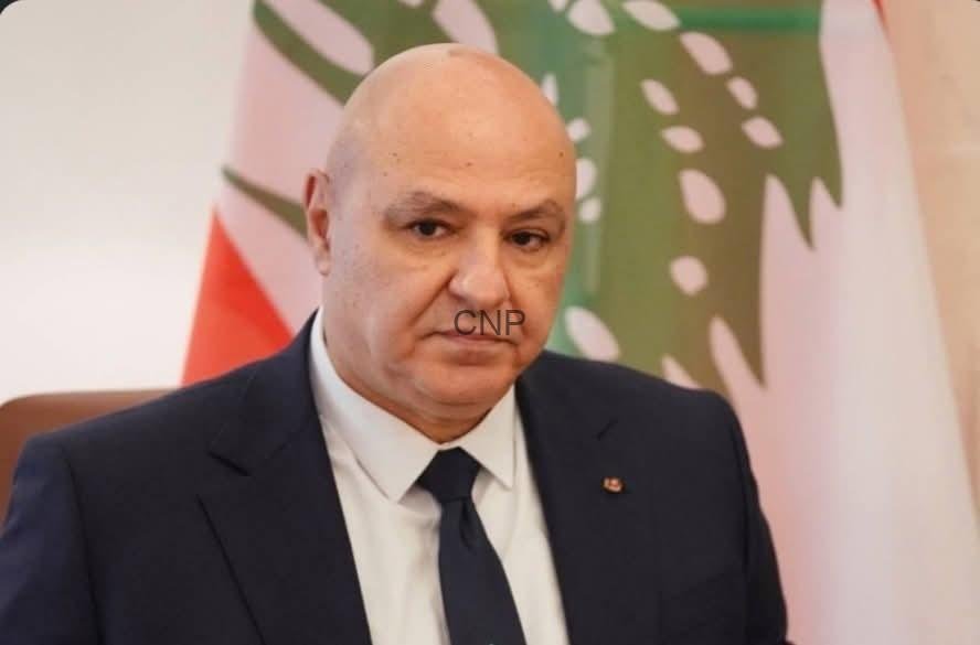 Bald man in a dark suit and tie sits at a formal event with flags in the background, looking toward the camera.