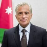 Formal portrait of a man in a dark suit and brown tie, with a Jordan flag in the background.