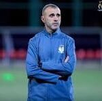 Man in blue sportswear standing with arms crossed on a soccer field.