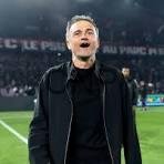 Soccer coach on the field celebrating with mouth open, wearing a black jacket, stadium lights in the background.