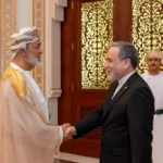 Two men shake hands in an ornate room; one in traditional Omani attire and the other in a dark suit, with a uniformed attendant in the background.