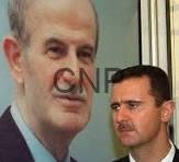 asad - Center News Network Two men in suits standing indoors in front of a large poster showing a man's portrait in the background.