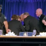 Security guards in tactical gear flank a conference table on a stage as suited people lean in for a discussion.