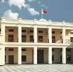 Two-story beige building with a row of columns, a flag on the roof, and a palm tree on the left under a blue sky.