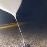 White helium balloon tied to a small rock on a sandy surface, under a dark night sky with a faint road line in the background.