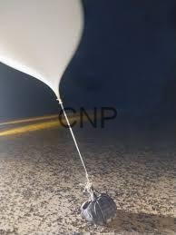 bal - Center News Network White helium balloon tied to a small rock on a sandy surface, under a dark night sky with a faint road line in the background.