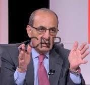balid - Center News Network Middle-aged man in a suit and tie, wearing a lapel microphone, gesturing while speaking on a TV interview set.