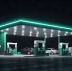 Nighttime gas station with green pumps and turquoise neon roof lights, a few cars parked at the islands.