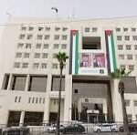 Facade of a large white multi-story office building with a wide entrance and a vertical green, white, and red stripe on the facade; cars parked along the street in front.
