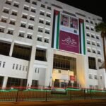 Night view of a white government building at night with a large banner featuring portraits and Arabic text, bordered by Jordanian flag colors and lights along the fence