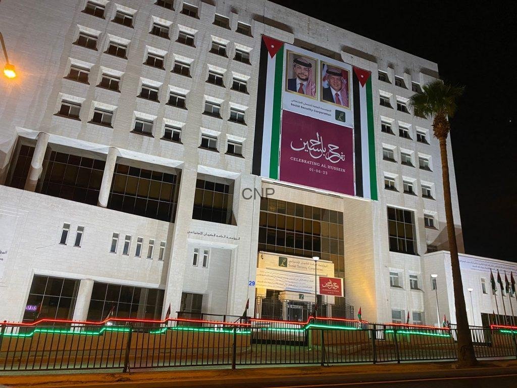 Night view of a white government building at night with a large banner featuring portraits and Arabic text, bordered by Jordanian flag colors and lights along the fence