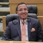 Man in a dark suit with a peach tie sits at a desk in a formal courtroom or council chamber.