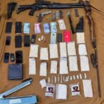Weapon and contraband stash: several firearms, magazines, and ammunition laid out with money and phones on a brown backdrop for display or evidence.