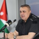 Man speaks at a press conference, seated at a table with a microphone in front, Jordan flag in the background.