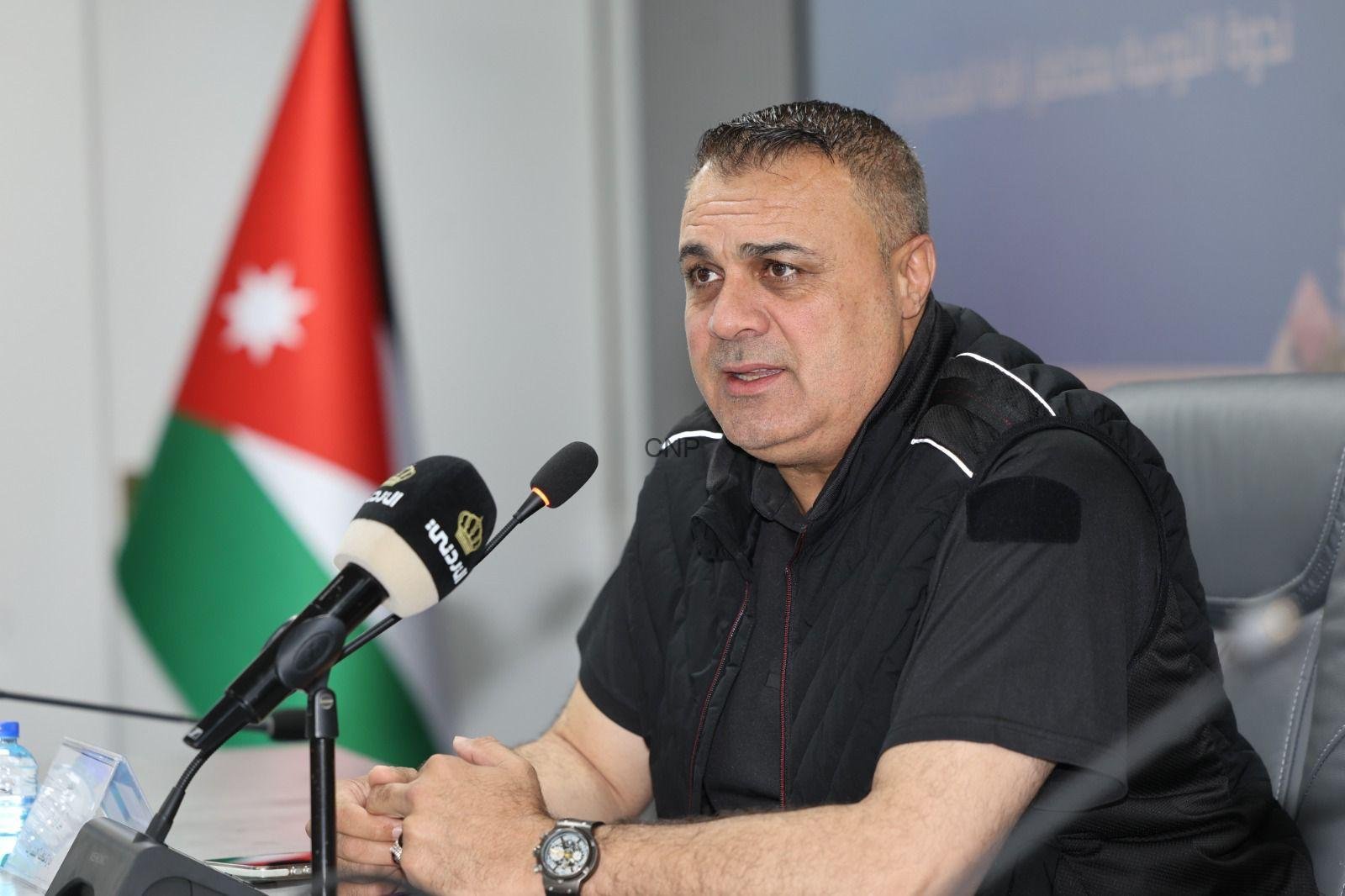 Man speaks at a press conference, seated at a table with a microphone in front, Jordan flag in the background.