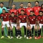 Eleven football players in red jerseys and black shorts pose for a team photo on the field, with a goalkeeper in teal at the left.