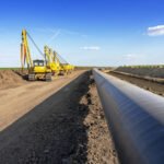 Yellow excavators lay a large underground pipeline along a dirt road under a blue sky, during construction.