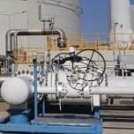 Industrial piping system with valves and gauges at an outdoor facility, mounted on blue supports under a clear sky.