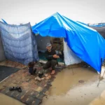 Man crouching at the entrance of a blue tent in a flooded refugee camp, a child nearby, with muddy ground and tarps around.