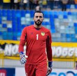 Male soccer goalkeeper in a red kit standing on the pitch with blue stadium seats in the background.