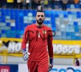 goa - Center News Network Male soccer goalkeeper in a red kit standing on the pitch with blue stadium seats in the background.