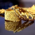 Close-up of ornate gold bracelets and rings on a mirrored surface, with floral engravings and reflections.