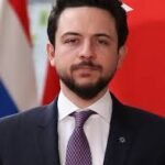 Professional portrait of a bearded man in a navy suit and purple tie, standing before red and white flags in the background.