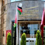 Front of an Arabic-language government building with gold signage, flags, and green shrubs in the foreground.