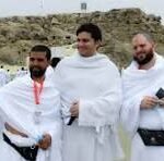 Three men in white robes pose together on a sandy beach during a ceremony, with onlookers and distant rocks in the background.
