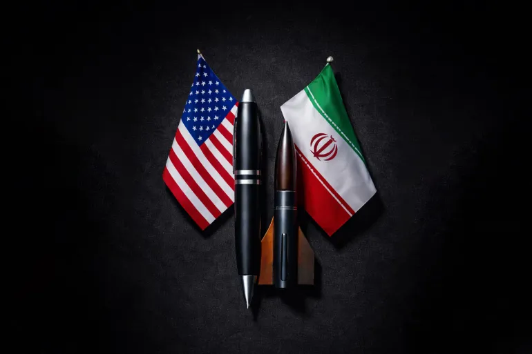 i u - Center News Network Flag of the United States and Iran displayed beside three pens on a dark surface.