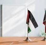 Two small national flags (Nigeria and Italy) stand on a wooden conference table in an office setting.