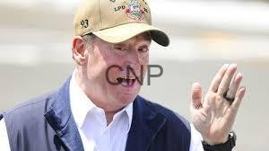 jone - Center News Network Man wearing a beige cap and blue vest waves with his right hand outdoors.