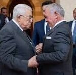 Two older men in dark suits shake hands in a doorway, with several suited bystanders watching.