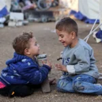 Two toddlers sit on the dirt and share a rock-and-block toy, smiling at each other near a campsite with tents in the background.