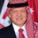 Portrait of a man wearing a red-and-white keffiyeh and dark suit, with flags blurred in the background.