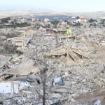 Rubble-strewn hillside town with collapsed buildings and debris, two people walking along a dirt path in the foreground.