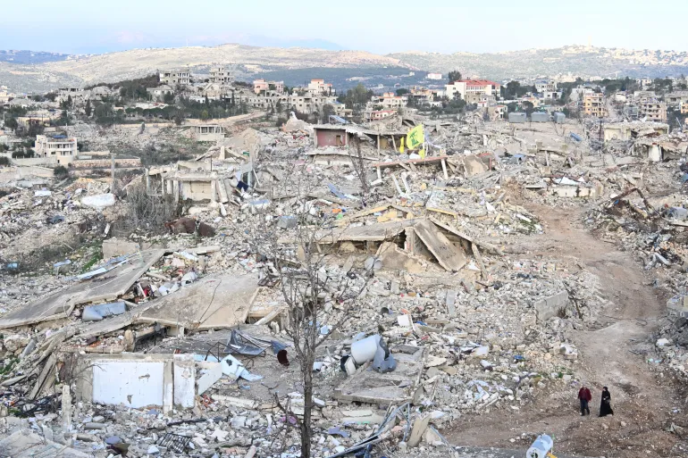 Rubble-strewn hillside town with collapsed buildings and debris, two people walking along a dirt path in the foreground.