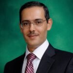 Professional head-and-shoulders portrait of a man in a dark suit, white shirt, and striped red tie, against a green background.