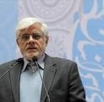 Older man with white hair and glasses in a dark suit speaks at a podium during a conference, with a blue patterned backdrop.