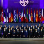 Group photo of world leaders at a NATO summit, with numerous national flags and the NATO emblem above them on a purple-lit backdrop.