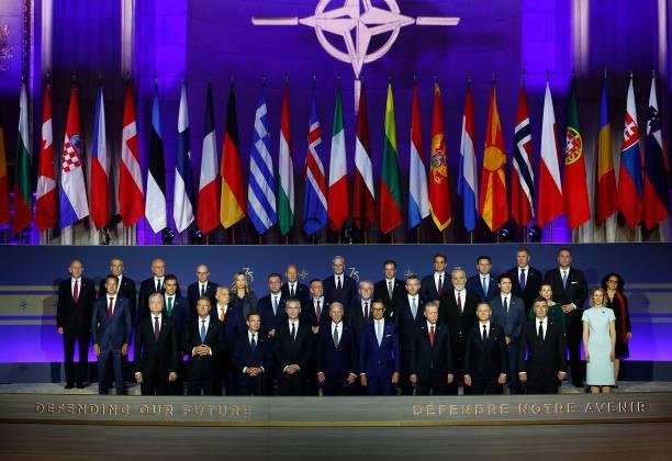 nato - Center News Network Group photo of world leaders at a NATO summit, with numerous national flags and the NATO emblem above them on a purple-lit backdrop.