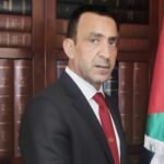 Man in a dark suit and red tie standing in front of a bookshelf with leather-bound books and a Jordanian flag nearby.