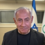 Older man in a dark blazer speaks to the camera, with flags in the background.