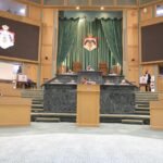 Legislative chamber with a central speaker's podium, green drapes, and crest emblems; officials seated at marble desks on a stepped dais.