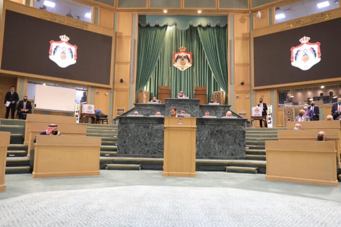 Legislative chamber with a central speaker's podium, green drapes, and crest emblems; officials seated at marble desks on a stepped dais.