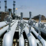 Rows of large metal pipelines with circular valve wheels at an oil refinery or processing plant.