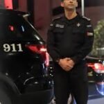 Uniformed police officer in black attire standing with hands clasped beside a marked police SUV showing '911' at night.